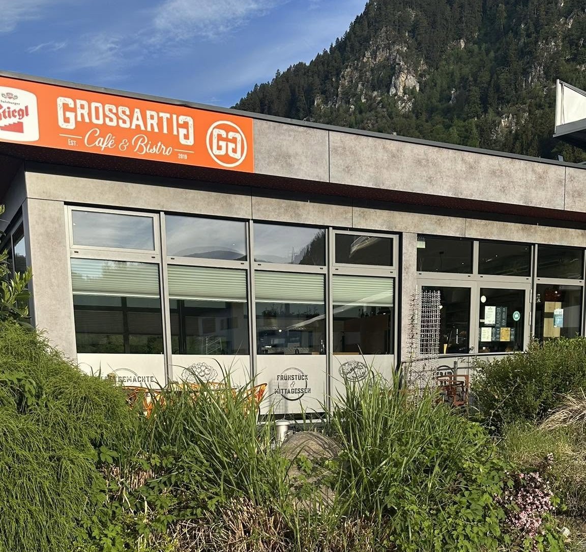 Außenansicht des Café & Bistro Grossartig mit orangem Leuchtschild, Terrasse und Bergen im Hintergrund